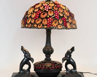 440	Art Deco Lamp With Richard Hoosin Shell Shade	Richard Hoosin (American, 1944-). Leaded shade with shells, stained glass and glass beads, signed Hoosin and dated 95 on the cap. On a later art deco style lamp, patinated metal with shells and glass beads, mounted on a slate base with a pair of patinated metal seal figures. Shade 14"-diameter, lamp with base and shade 19 1/2"L x 10"W x 24"H including finial. Slate base missing foot, chip along back edge of slate, lamp not attached to base.
