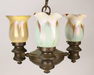 229	Brass Chandelier With Quezal Shades	Brass hanging fixture, with 3 iridescent art glass pulled feather shades. 2 signed Quezal, 1 mismatched shade, unsigned. Fixture with shades 9 3/8"L from top of ring (not including chain and cap), 11 3/4"-diameter. Quezal shades 4 1/2"-diameter, fitter 2 1/8"-diameter. One quezal shade chipped at rim of fitter, one arm missing screw for shade, paint spots and losses to brass patina on fixture.
