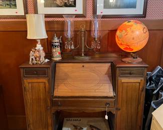 This photo shows several items, including the writing desk, a lighted globe, a candelabra, beer stein, framed artworks, and tabletop lamp. 