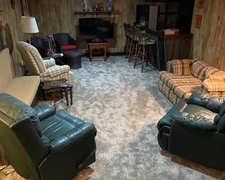 Throw back basement rec room offers a plaid love seat, two leather recliners, an upholstered recliner, Sony flat screen TV,  plastic wicker patio chairs with ottoman and side table, swivel bar stools and lots of misc