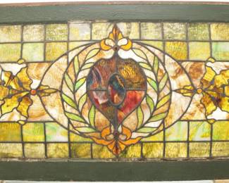 A turn of the century American stained glass window.  Curved window with multi-color stained and leaded glass panels depicting a scrolling Heraldic design with faceted and textured glass accents.  A few cracked panels with losses to glass.  In a painted wooden frame with wear, 42 1/2 x 20 1/2" high overall.