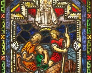 A Gothic Revival stained glass window of Our Lady of Charity.  Multi-color stained and leaded glass panels with reverse painted detail depicting imperiled sailors praying to the Holy Mother and Child within geometric and foliate borders.  One cracked panel at oar, some wear to paint.  In a later Oak frame, 23 x 32" high overall.