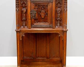 A turn of the century Gothic Revival Period carved Oak court cupboard.  Molded top over one central door with carved tracery and shield, flanked by carved figural panels and spires, over an open lower shelf with linen fold carved back panel, Iron hinges and figural Apostle lock.  Original finish with some wear and damage, glued repair to lower carved skirt.  34 3/4 x 17 1/2 x 56 1/4" high overall.