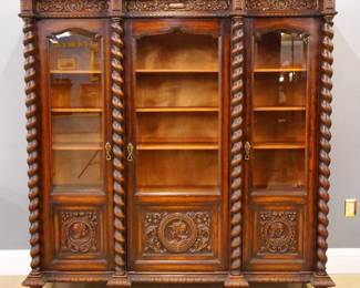 A turn of the century Gothic Revival Period Oak bookcase.  Molded top over a carved frieze and three doors with beveled glass panes and carved portrait lower panels, flanked by barley twist columns on a carved bracket base with six turned feet.  Older dark finish with some wear, minor damage.  75 x 19 3/4 x 76" high.
