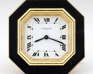 A 1970s Cartier travel alarm clock.  Manual wind Swiss movement with painted metal dial and Roman numerals.  Octagonal Gilded Brass case with Black enamel accents and hinged stand with Cartier "C" feet.  Impressed "Cartier Paris" and "7507 02489 / Swiss Made" at back.  Some wear to enamel, running when cataloged.  3 1/8" high.