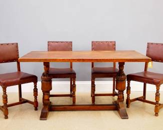 A turn of the century Jacobean Revival Oak dining set.  Dovetailed plank top table on a trestle base with two turned pedastal legs with carved detail.  Four side chairs with leather upholstered backs and seats on stretcher bases with turned front legs.  Old finish with wear and minor shrinkage.  Table is 58 3/4 x 29 3/4 x 30 1/2" high overall, chairs each 34 3/4" high.