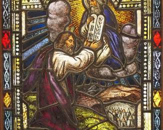 A Gothic Revival stained glass window of The Ten Commandments.  Multi-color stained and leaded glass panels with reverse painted detail depicting the Moses receiving the Ten Commandments from God within geometric and foliate borders.  One panel cracked below Moses' knee, three panels at pediment with restored finishes, some wear to paint.  In a later Oak frame, 20 1/4 x 34 1/4" high overall.
