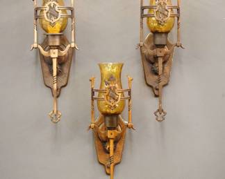 A set of three of early 20th century Gothic Revival Period sconces.  Cast Bronze bodies with Shield design and patinated finish, single lights with Amber crackle glass hurricane shades.  Some wear to patina and paint splatters, glass shades with some chips and flakes.  Each 16 1/2" high.