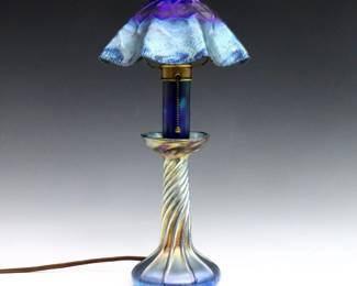An early 20th century Tiffany Favrile Glass candlestick lamp.  Ruffled Iridescent Blue glass shade with on candlestick base with swirled design.  Shade signed "LCT" and base "L.C.T / Favrile".  Minor surface wear, replaced Tiffany shade with flake to edge.  7" diameter x 13 3/4" high overall.  
