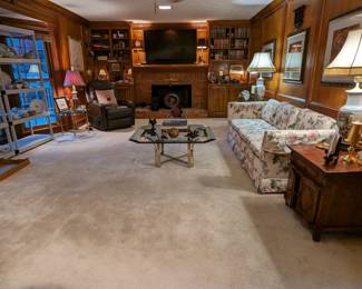 Long shot of the comfy den with a pair of hand-carved wooden side tables with single drawer and two doors, pair of Asian porcelain table lamps, brass/beveled edge glass coffee table with stack of good books and hand-carved wooden fish collection, pair of genuine leather power reclining/lift chairs, pair of round brass/glass side tables with pair of matching glass/resin table lamps, built-in bookshelves with nice collection of automobile books, cast iron toys, travel souvenirs, etc.