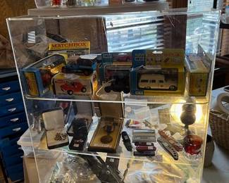 This showcase contains Matchbox cars and trucks in original boxes in great condition, WWII military medals, pocket knives, large hunting knife, vintage belt buckle, Harley Davidson patches and Hog pins. Designer perfume