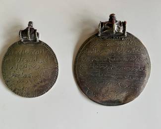 A pair of Omani silver Samt pendants with text from the Quran on the front and a shackled jinn on the reverse—these were worn to ward off bad luck