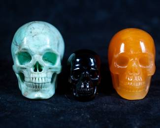 Three carved crystal skulls: One made of Orange Calcite, One made of Smokey Quartz, and One made of Picasso Jasper.
