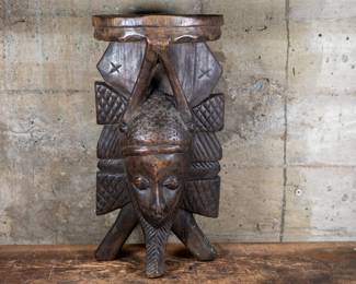 A wood carved mask originating from Gambia. The mask is designed to stand on a table with a small shelf on the top.
