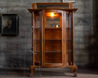 A Mid-Century Curio Cabinet made of Oak, made by Holman with three shelves, a top with rails and a mirror back with wooden claw feet and an interior light.