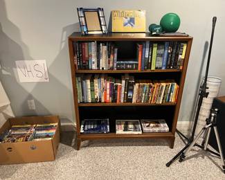 Lower Level:  A box of VHS tapes is to the left of a bookcase filled with books.  To the right are a SAMSON microphone stand with boom arm and tripod base AND a HOLLYWOOD PC-3 camera tripod.