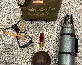 Lower Level:  A vintage zipper lock bank bag (The  Union National Bank, Springfield, MO) is near a sling shot; a bullet-look flashlight; an antique iron shot put ball; and a circa 1970 STANLEY "Aladdin" stainless steel green vacuum thermos.