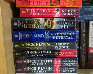 Military And Espionage Paperback Books Including Robert Ludlum, Vince Flynn, Ken Follett, And More, Contents Of 3 Shelves