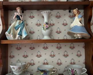 A shelf brimming with ceramic woman figurines that are planters and vases, vintage cups and saucers, a Lefton angel for March and other delightful finds. 