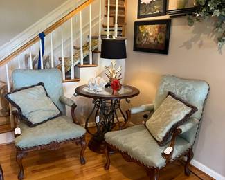 Green upholstered armchairs with pillows, round wood/ glass/iron table, lamps, art