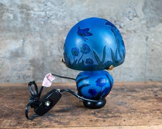 A blue glass mushroom-shaped lamp with garden designs. The lamp has multiple light modes, with lights in the base of the lamp and at the top, with a mode where the light occasionally flickers at the top to simulate a firefly. Tested and in working condition.