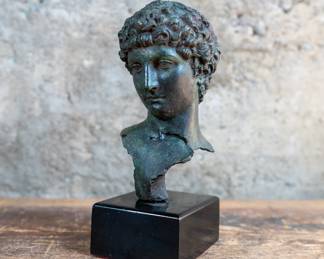 A bust of a Greek man, with curly hair and a serene facial expression. Constructed out of grey polymer on a black wooden plinth.