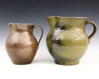 Two late 20th century Jugtown Pottery pitchers.  Bulbous forms with ruffled rims and strap handles, includes a Ben Owen pitcher with mottled Green Frogskin glaze, and a Vernon Owens pitcher with Brown salt glaze.  One with stamped "Ben Owen / Master Potter" mark, the other with Impressed "Jugtown Ware" mark, "1982" date and incised "Vernon Owens" signature.  Minor wear, Ben Owen pitcher with chip to rim and larger burst air bubble from production at body.  7" and 8 1/2" high.  ESTIMATE $50-100