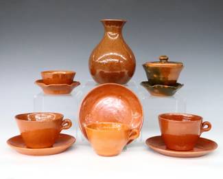 Nine pieces of 20th century Jugtown Pottery.  Various forms with mottled Orange, Orange/Green Accidental and Red/Brown Tobacco Spit glazes.  Includes a vase, covered bowl with incorporated ruffled underplate, three teacups and three saucers, and a candle holder with incorporated ruffled underplate.  Some with crazing, minor surface wear and flakes.  Up to 5 3/4" diameter and 6" high.  ESTIMATE $50-75