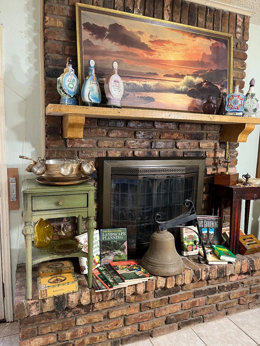 Notice the Jim Bean whiskey decanters on the mantle by the vintage sea scape. On the hearth is a vintage small table with a brass punch bowl set on it. Surrounded by vintage gardening books is a magnificent large brass bell.