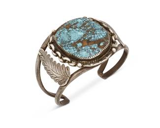 387
A Navajo Sterling Silver And Set Turquoise Cuff Bracelet
Mid/late 20th century
Stamped: JAG [conjoined, with serifs] / Sterling
A two wire cuff with central slab turquoise with heavy matrix and applied feather elements to shoulders
6.6" inner C x 1.75" H, with a 1.125" gap
43.0 grams gross
Estimate: $700 - $900
