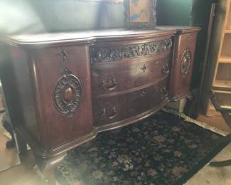 Large oak buffet, beautiful relief work and carving. Black floral wool rug. Vintage tin relief photo frame with wood backing and stand atop buffet.