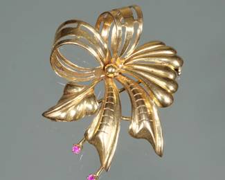 18K GOLD RIBBON FORM PIN | Designed as an 18 karat gold pierced open work, ribbon form pin with a leaf and mounting two rubies; 2 in., 10.2g; with maker's marks and fineness marks.
