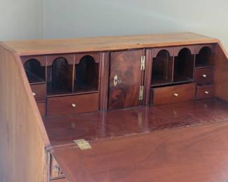 Closeup of antique slant front desk, top opened as writing surface, resting on supports; showing interior drawers, cubby holes, and additional locked compartment