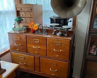 In this photo, you can see the five-drawer filing cabinet.  Two of the drawers are extra wide, probably for legal-sized documents.  On top, the lockable four-drawer card catalog with a sliding front door.  On the right, an Ohaus balance scale, sitting in front of the vintage record player with bell horn.  Could be a Victrola, but possible an Edison or another maker's?