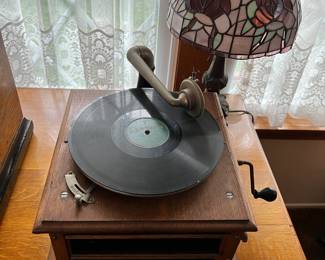 This hand-crank record player has a Columbia Grafanola, sitting in front of a Tiffany-inspired table lamp.