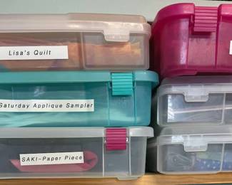 Multiple plastic boxes each with a quilt project (instructions, some fabric, often completed pieces)