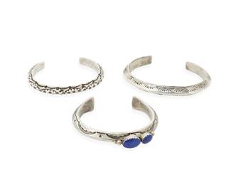 293
Three Southwest silver cuffs
Late 20th/21st century
Three works:

Jeanette Dale (b. 1949; Diné)
Stamped: JDale [conjoined] / Sterling
A narrow Navajo ridged sterling silver cuff with set lapis lazuli cabochons and elaborate stampwork, with flared terminals
6 1/2" inner C x 5/8" H, with a 1 1/4" gap

Two sterling silver cuffs
Each unmarked
Each narrow ridged cuff with elaborate stampwork
Larger: 7" inner C x 3/8" H; smaller: 6 1/2" inner C x 5/16" H

3 pieces total
149.4 grams gross
Estimate: $200 - $400