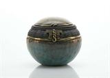 A pretty hand crafted round pottery jar with separate dome shaped lid, decorated in a teal green with coppery luster finish and a band of black to the top of the jar which has a basket weave texture and applied gold accents. Seemingly signed “Caroline” to the bottom.