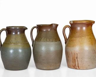 Three Salt-Glazed Stoneware Pitchers, Middle TN origin, late 19th or early 20th century, all of similar ovoid form, the two smaller examples with incised wavy line decoration to base of collar, each dipped in brown slip and covered in a clear salt glaze. Tallest with a 1" rim chip. Middle-sized pitcher with a 1/2" rim chip, a minor chip to spout, and a small in-the-firing surface flake to body. Smallest with shallow chips and wear to rim and spout, a hairline through handle, and surface wear to body, rim, and handle, the handle with minor traces of old paint spatter. H (of tallest) 11 1/4". ($200-$400)
