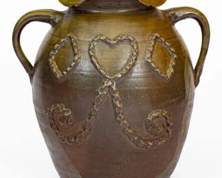 Exceptional Salt-Glazed Stoneware Jar with Applied Decoration, attributed to the Hedgecough Pottery, Putnam County, TN, late 19th or early 20th century, ovoid jar with crimped rim and two vertical, open handles applied at the shoulder, decorated on the front with applied heart, diamond, and scrolled drape designs, all embellished with depressed treatment throughout. Surface dipped in a reddish-brown slip and covered in a salt glaze. One of a small group of documented jars decorated in this manner, this work ranks among the finest examples of brown-dipped Middle Tennessee stoneware known. Losses to rim. A reglued break to midsection of one handle. Otherwise excellent condition. H 13 1/2". ($3000-$5000)
