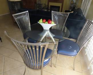 Small round glass table with brushed nickel legs and matching heavy metal chairs with blue vinyl seats.