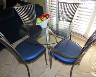 Small round glass table with brushed nickel legs and matching heavy metal chairs with blue vinyl seats.