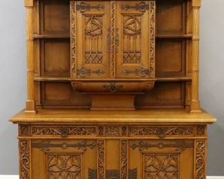 A turn of the century Gothic Revival Period sideboard.  Oak construction with extensive hand carved decoration and original embossed Iron scrolled hinge plates and escutcheons.  Features an upper section with a stepped rampart crest and foliate detail over two central doors with tracery panels flanked by open display shelves and carved columns on either side over a short central drawer, on a base with a molded top over two dovetailed drawers and two lower doors with tracery panels framed by carved foliate rails.  Older refinishing with some wear, minor damage, lacks some small moldings, base reduced in height approximately 2".  71 x 31 x 96 1/2" high overall.