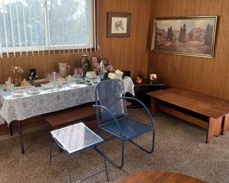 In this room, a pair of wooden benches sit underneath the framed artwork on the wall.  A glass side table and metal chair sit in front of a tableful of other treasures.