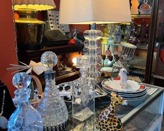CRYSTAL DECANTERS, HIGH-END LUCITE TABLE LAMPS, CHRISTMAS / HOLIDAY PLATE SETS... (IN BACKGROUND IS A BEAUTIFUL BRASS 1970'S ERA TABLE LAMP) ... AND A SMALL WOODEN DECOR ITEM: "SHELF GIRAFFE."