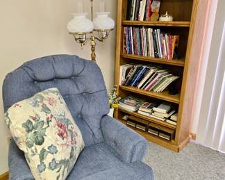 Blue Upholstered Casual Chair; Decorative Pillow; Double Milk Glass Shade Floor Lamp; Collection of Buttons in Jars; Oak Wood Tall Bookshelf; Cassettes; Books; Mantle Clock