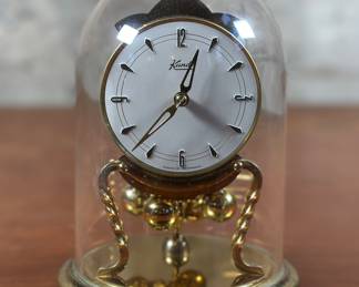 An alluring Kundo torsion clock, made in Germany in complete condition with original glass dome and key. This vintage mantle clock features an original white dial & matching twisted brass pillars, circa 1970s. This clock measures 6.5” H x 5” W x 5” D.