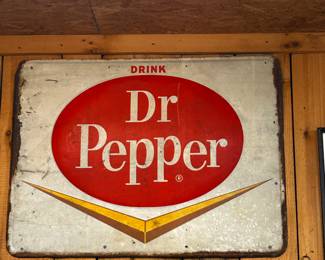 This is a vintage Dr Pepper advertising sign, likely dating from the mid-20th century. The sign features the classic Dr Pepper logo: a red oval with the words "Dr Pepper" in white, above a yellow and brown pointed design. The sign is made of metal, showing signs of wear and age, including rust along the edges and minor surface imperfections. The color palette and design elements suggest a production era between the 1950s and 1960s, a period when Dr Pepper's marketing heavily utilized this particular logo style. The sign's dimensions appear to be approximately 36 inches wide and 24 inches high, though this is an estimation based on the image. The sign would have been displayed in a retail environment, such as a soda fountain, grocery store, or gas station, to promote Dr Pepper sales.