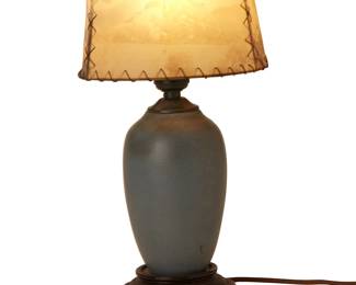 1061
Mid/late 20th century; Colorado Springs, CO
A Van Briggle pottery boudoir lamp
Appears unmarked
The single-light lamp with blue-glazed ceramic body on a wood base, surmounted by original hand-stitched parchment shade depicting cattle and flowers in a desert landscape, electrified
Overall: 13.5" H x 7" Dia.
Estimate: $400 - $600