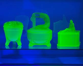 Blacklight photo of the vintage LE Smith uranium glass Greek key footed flower frog or Flowerlite vase, a Dermay Inc satin finish uranium glass Rapunzel powder jar, and a circa 1929 Phoenix Glass Co. frosted finish uranium glass powder jar
